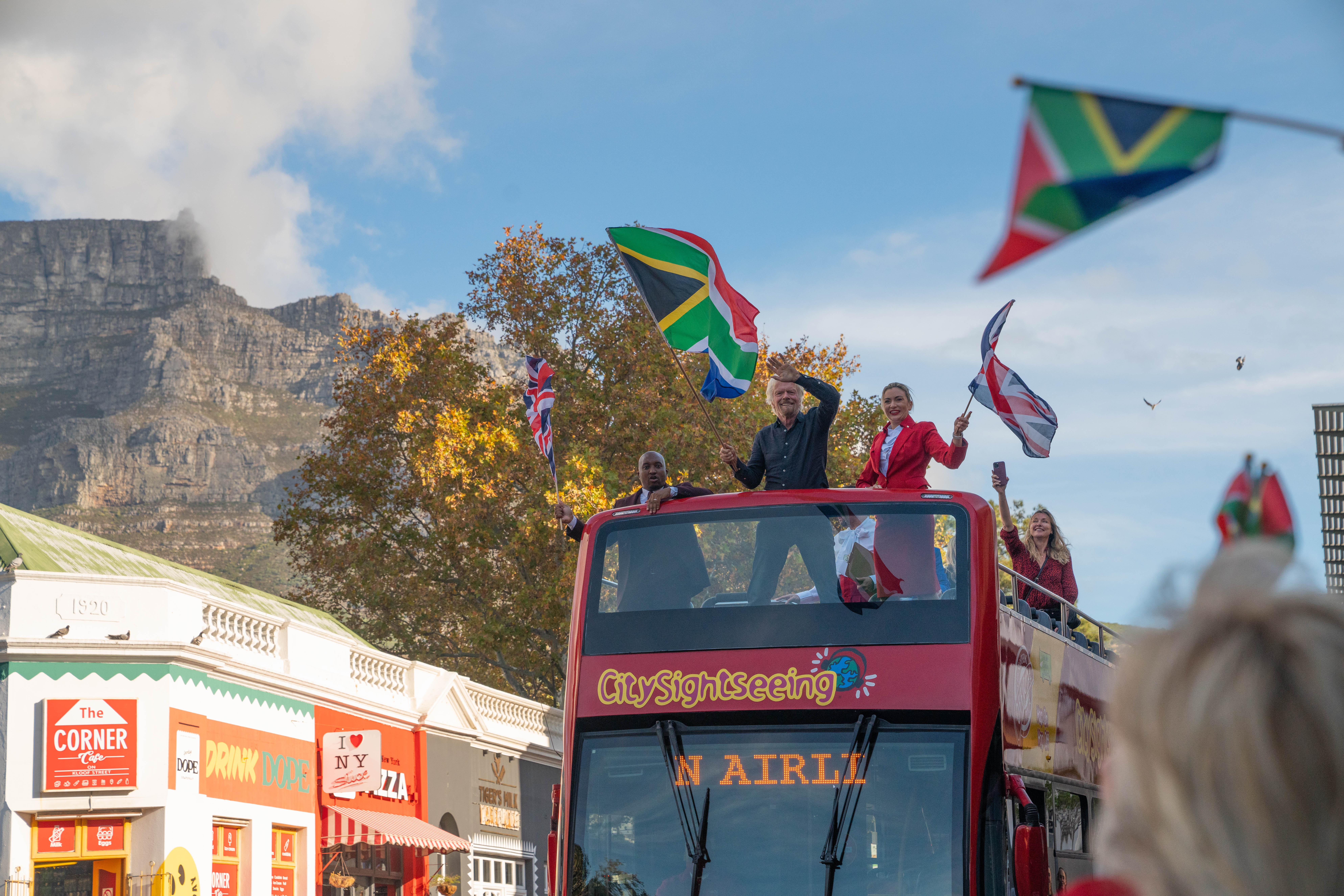 Photo of Richard Branson and two cabin crew on the top deck of a double decker tour bus raising flags for South Africa and the United Kingdom