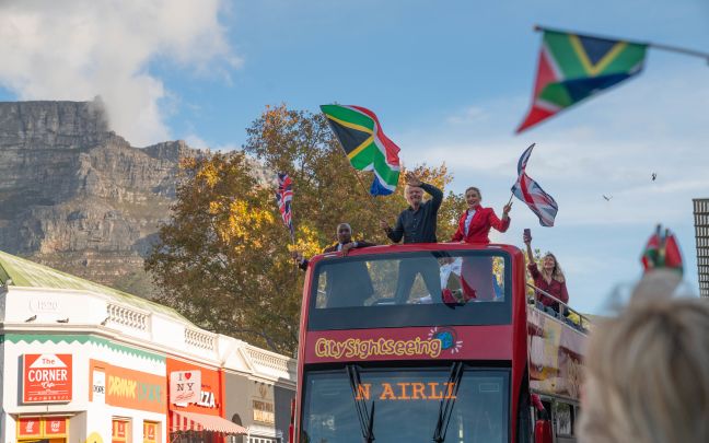 Photo of Richard Branson and two cabin crew on the top deck of a double decker tour bus raising flags for South Africa and the United Kingdom