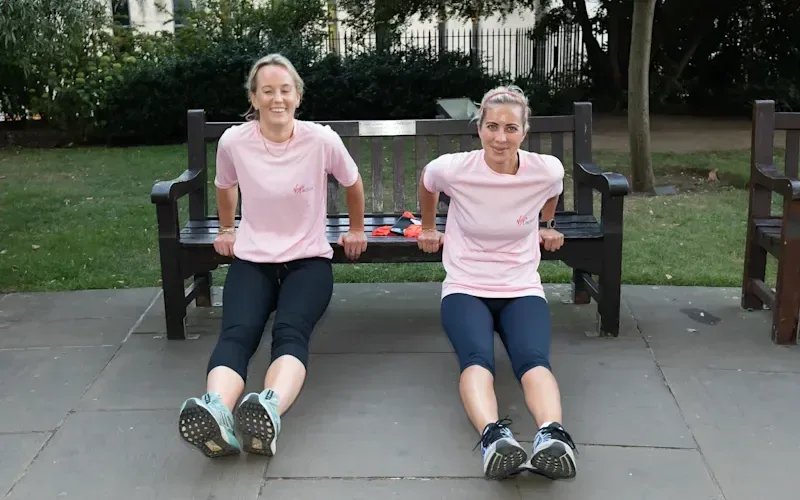 Holly Branson bench presses on a park bench with another woman, smiling at the camera