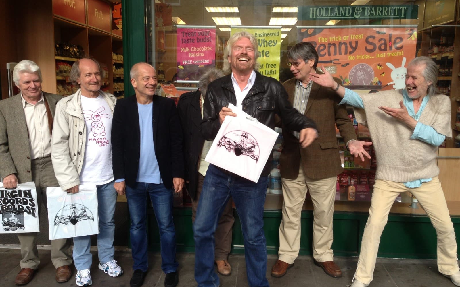 Richard Branson smiling with 6 other men laughing and standing in front of a shop