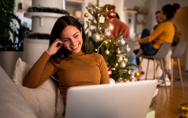 A woman sitting on the sofa using her laptop in front of a Christmas tree while other people sit on stools at a table in the background