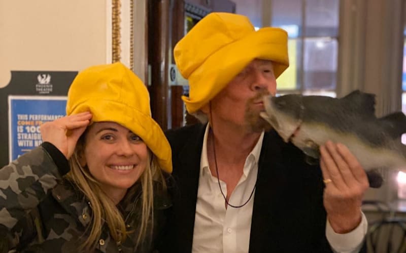 Holly and Richard Branson wearing yellow fisherman hats and Richard is kissing a plastic fish