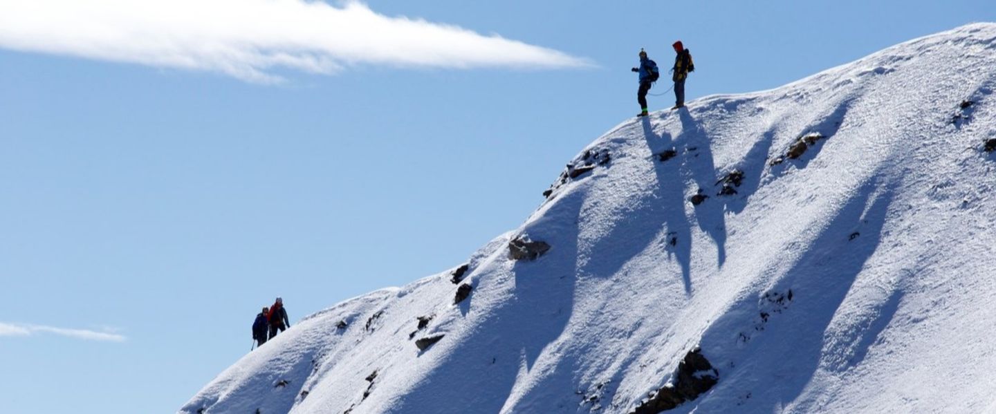 Two people stand at the top of a steep, snow-covered mountain