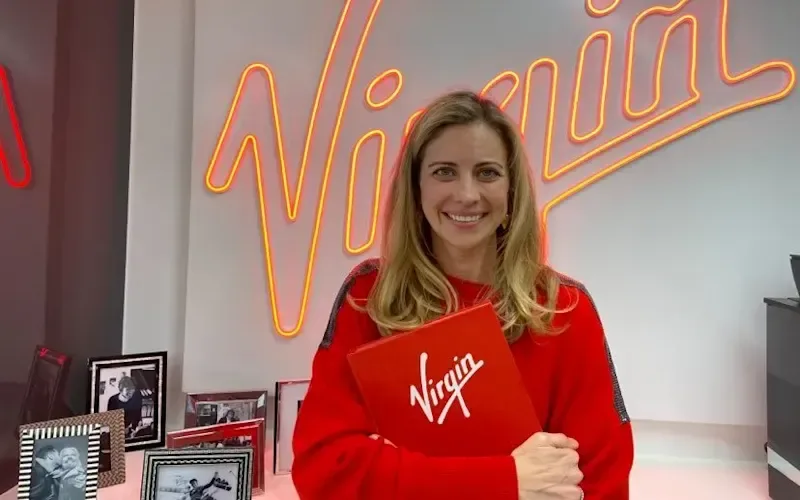 Holly Branson smiling, standing in front of a neon Virgin sign, holding a Virgin By Design book