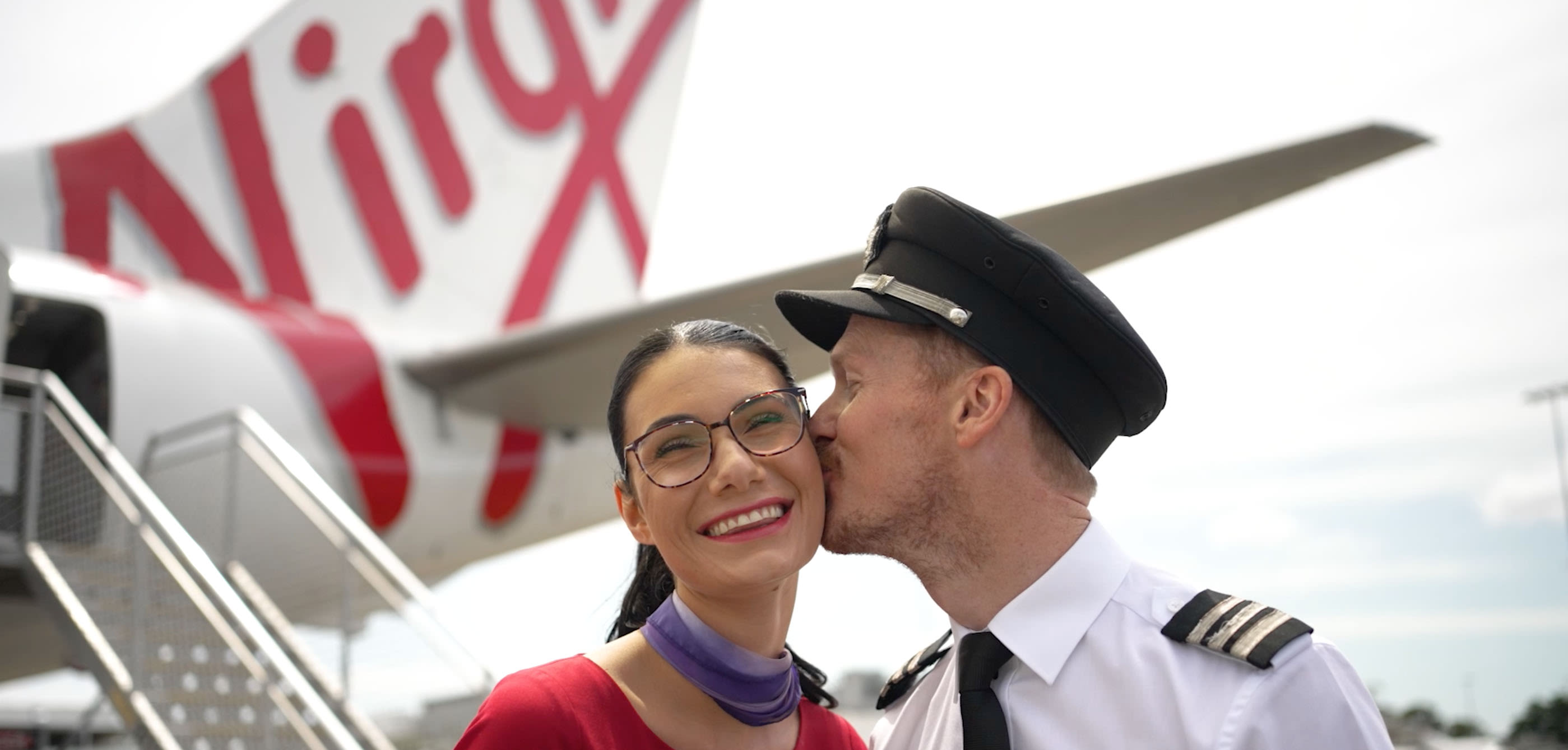 A Virgin Australia pilot kisses his cabin crew partner on her cheek in front of a Virgin Australia aircraft