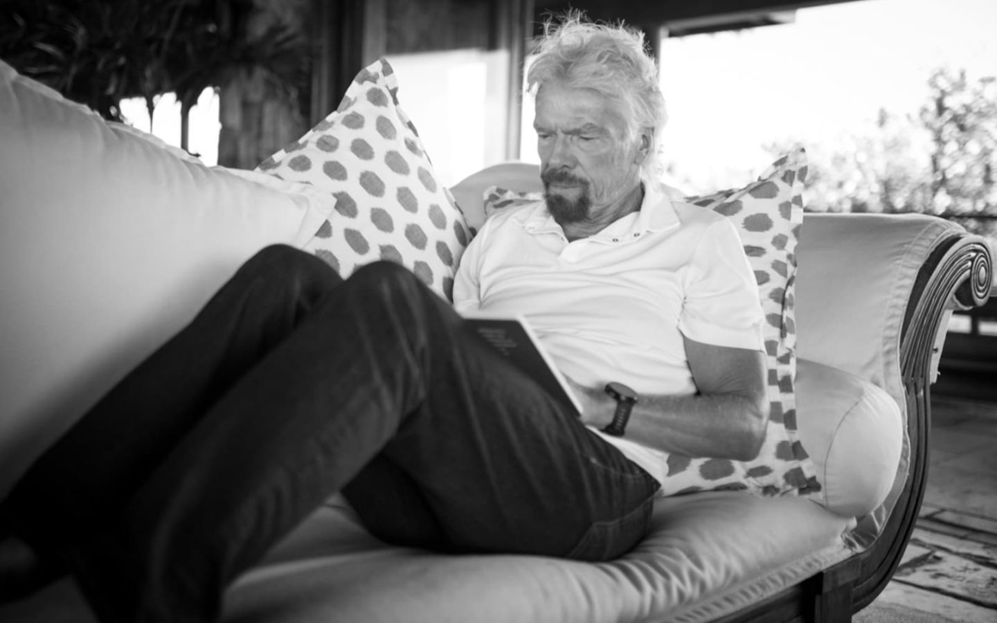 A black and white image of Richard Branson with his feet up on the sofa reading a book
