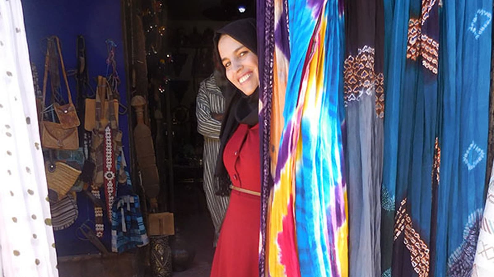 Woman wearing hijab and red dress smiling behind a colourful cloth inside her shop