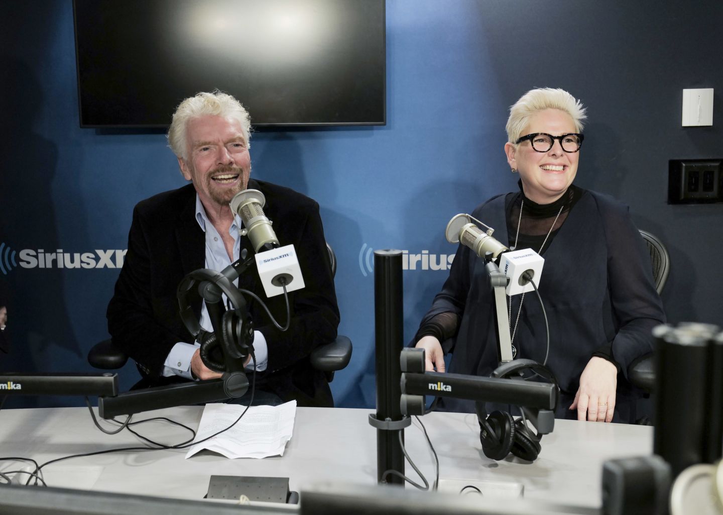 Richard Branson and Halla Tomas sitting behind microphones in the SiriusXM studio, smiling at the camera