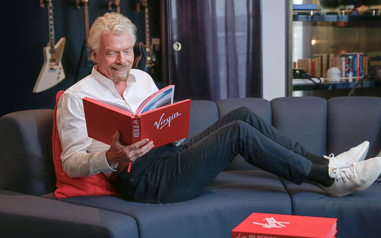 Richard Branson relaxing on a sofa reading a copy of Virgin By Design