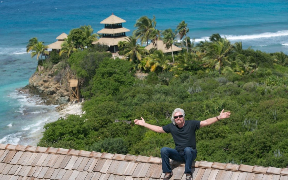 Richard Branson sitting on top of a roof with his arms stretched wide with the sea, forest add building from Necker Island in the background