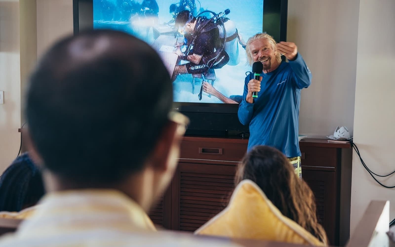Richard Branson in blue shirt speaking into mic in front of a TV