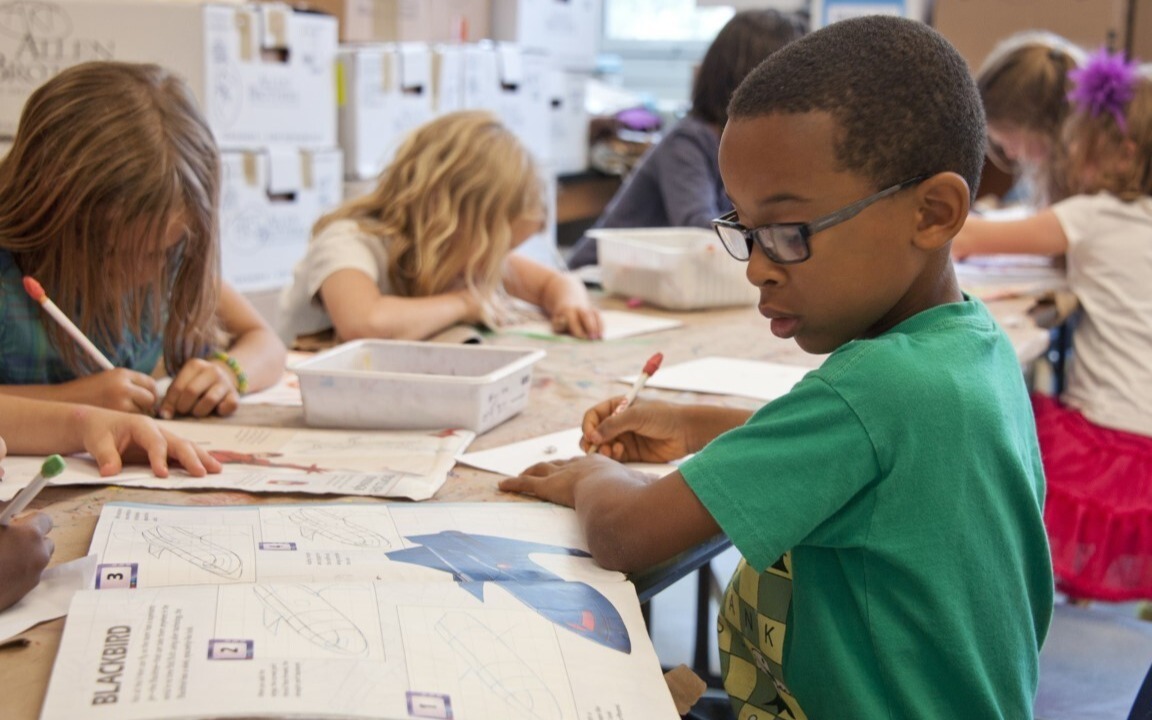 Image of a young boy working through a workbook at a table with other young children also writing.