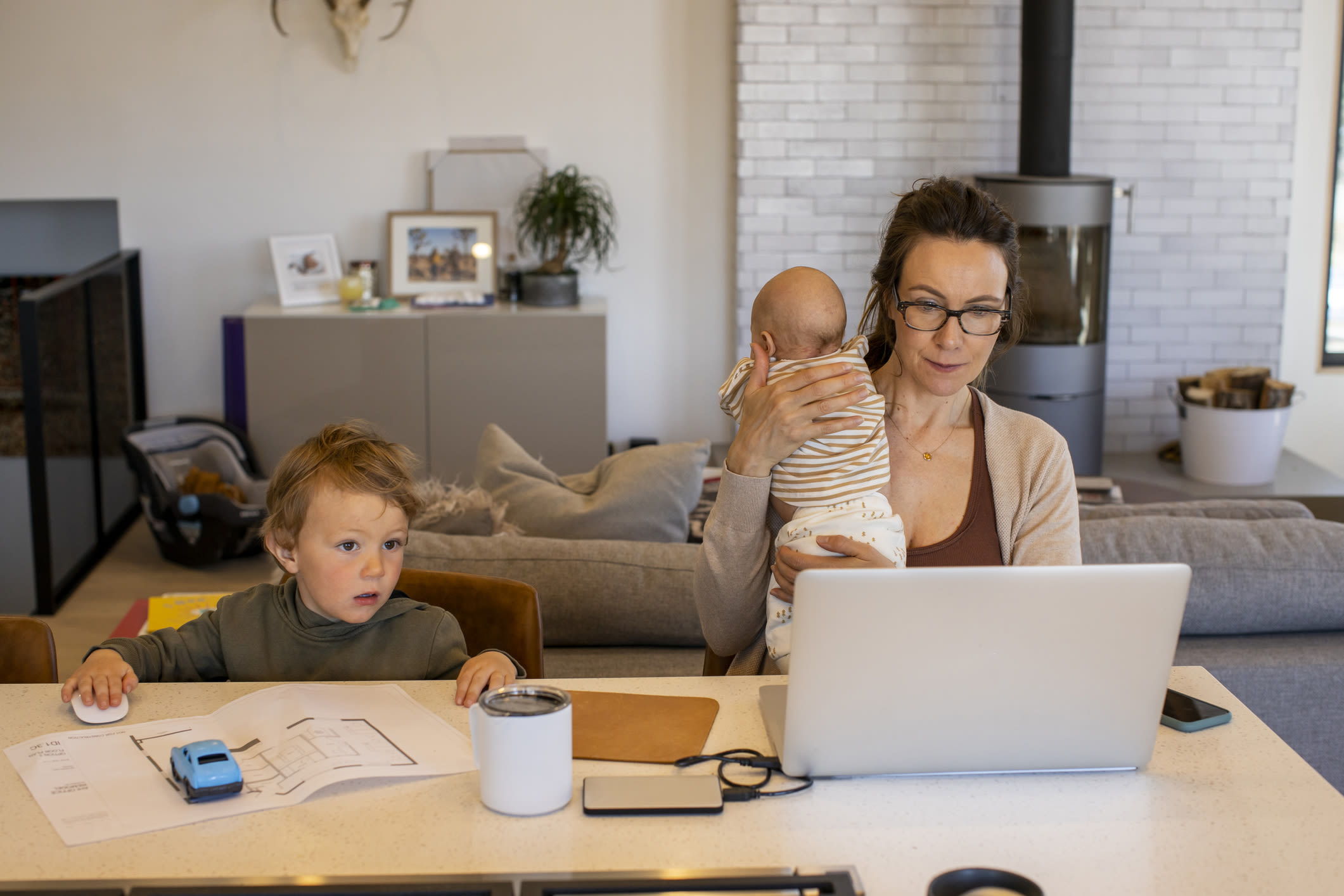 A mum working from home, holding a baby while an older child sits next to her at a table