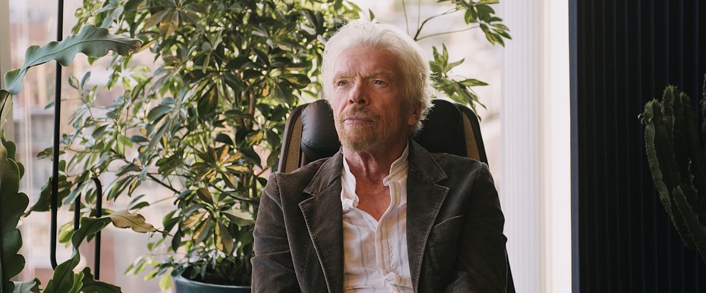 Richard Branson sitting in a chair with plants behind him looking serious as he looks off camera