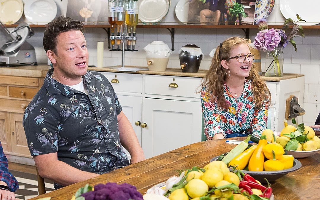 Jamie Oliver sat next to a lady smiling with fruit and veg on table in front of them