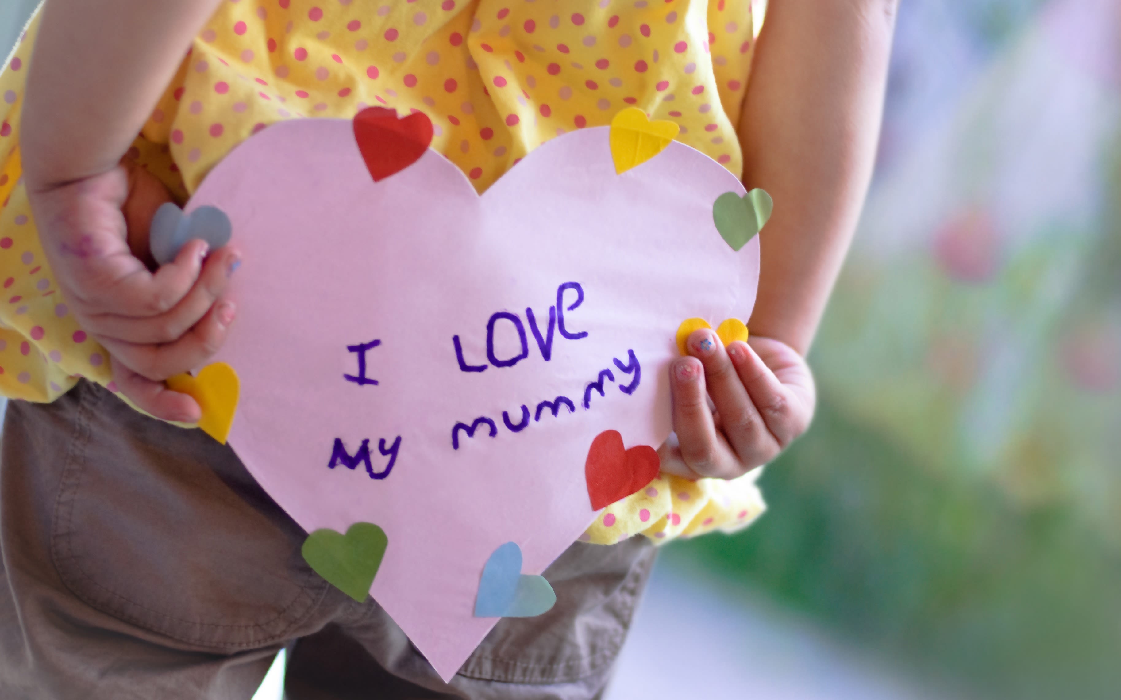 A child holding a heart-shaped sign that reads "I love my mummy" behind their back