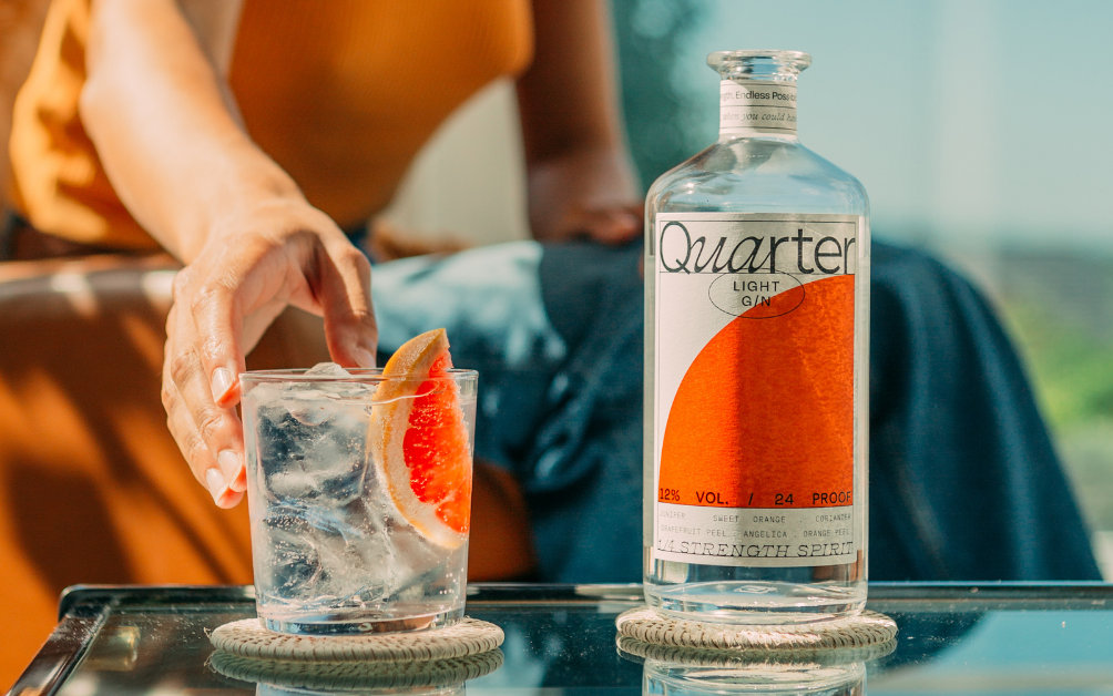 A person reaches for a glass with a clear drink and a slice of orange, next to a bottle of Quarter