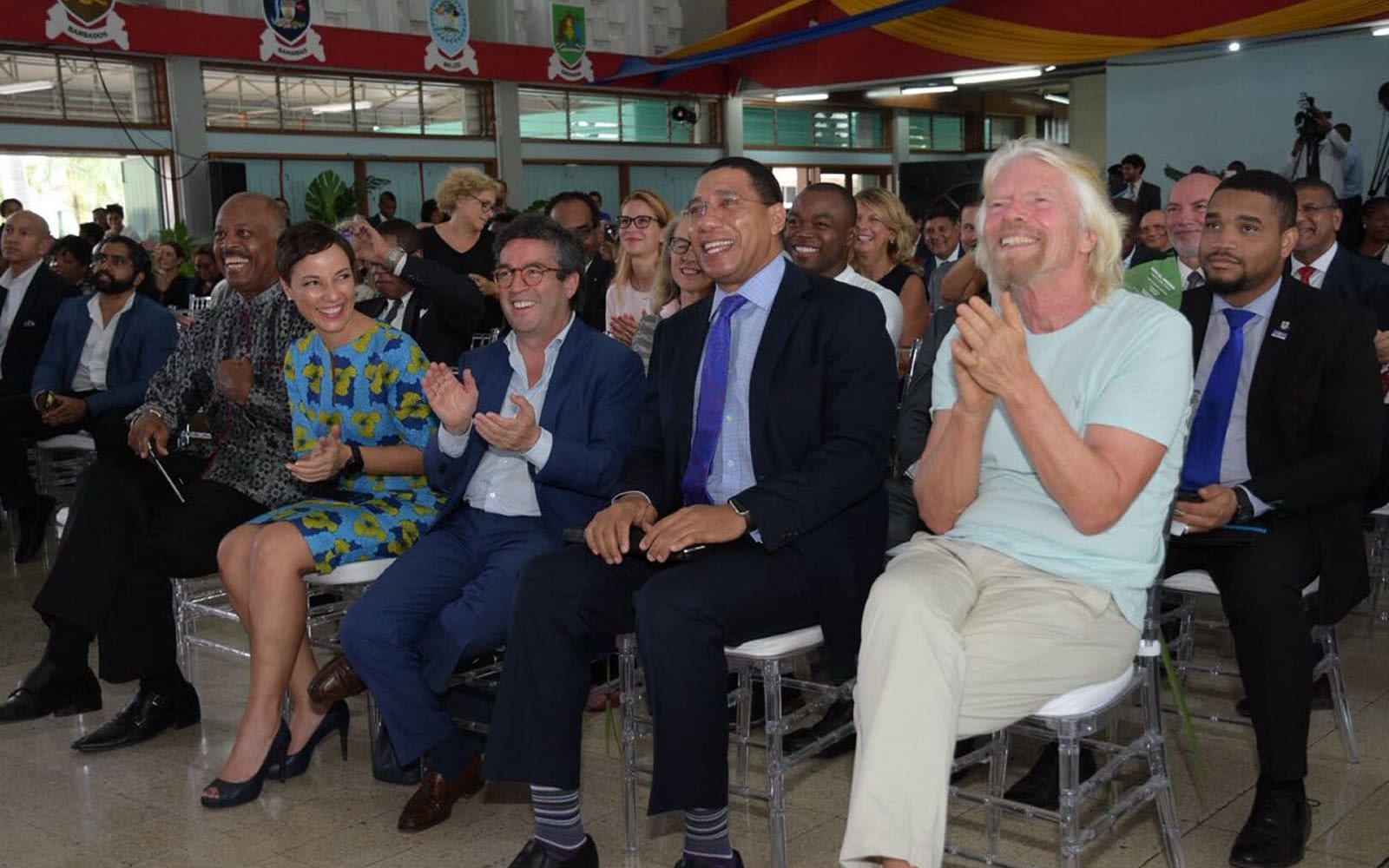 Richard Branson wearing a light tshirt and sat down with guests at an event smiling and clapping 