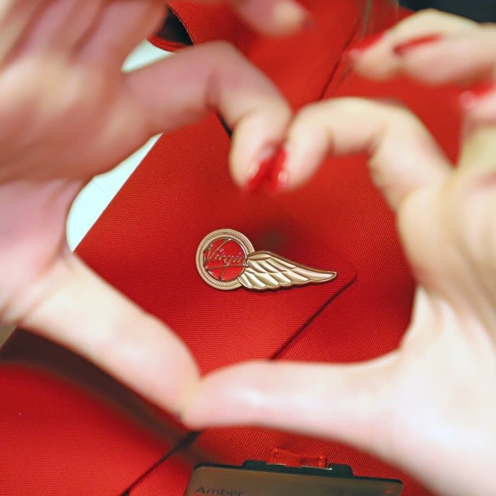 A woman's hand making a love heart shape over the Virgin Atlantic cabin crew wings pin on her cabin crew uniform