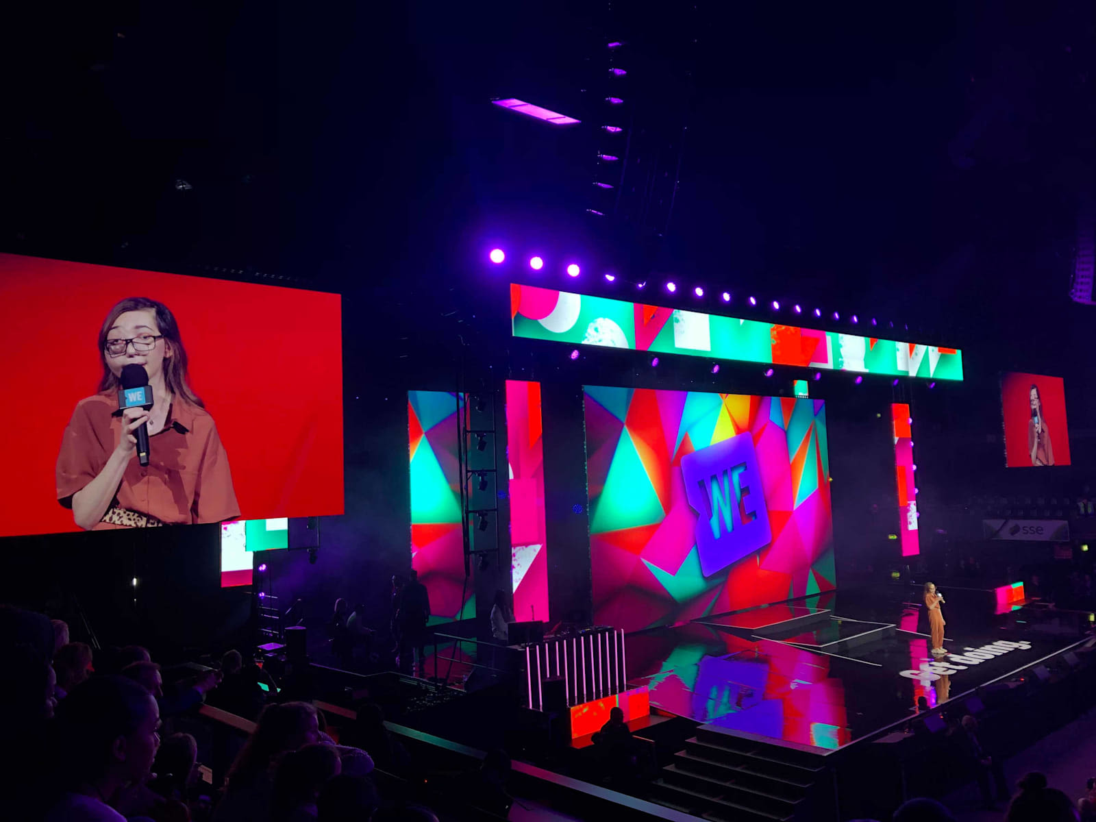 Picture of the stage from the left again with multicoloured lighting and a big screen to the left showing a woman talking to the crowd into a microphone 