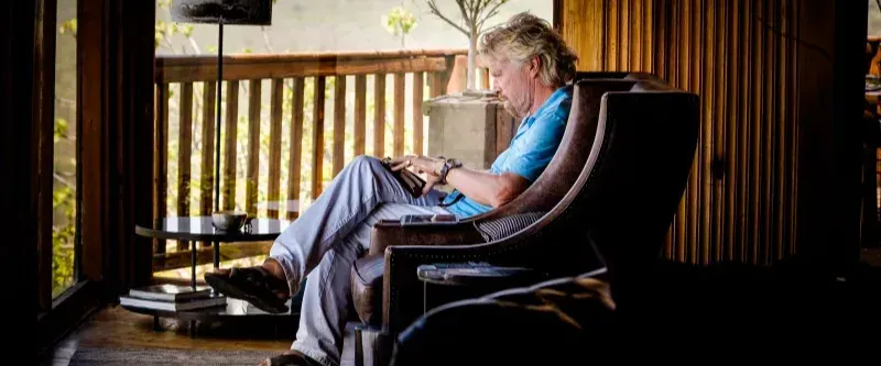 Richard Branson.  Side profile, sat in a chair writing notes