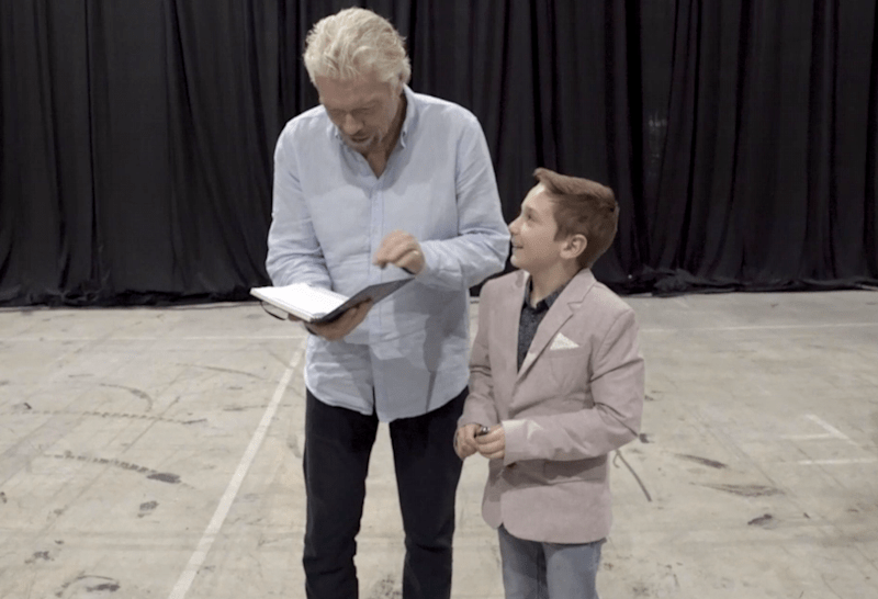 Richard Branson looking at his notebook. He is stood next to a child who is looking up at him