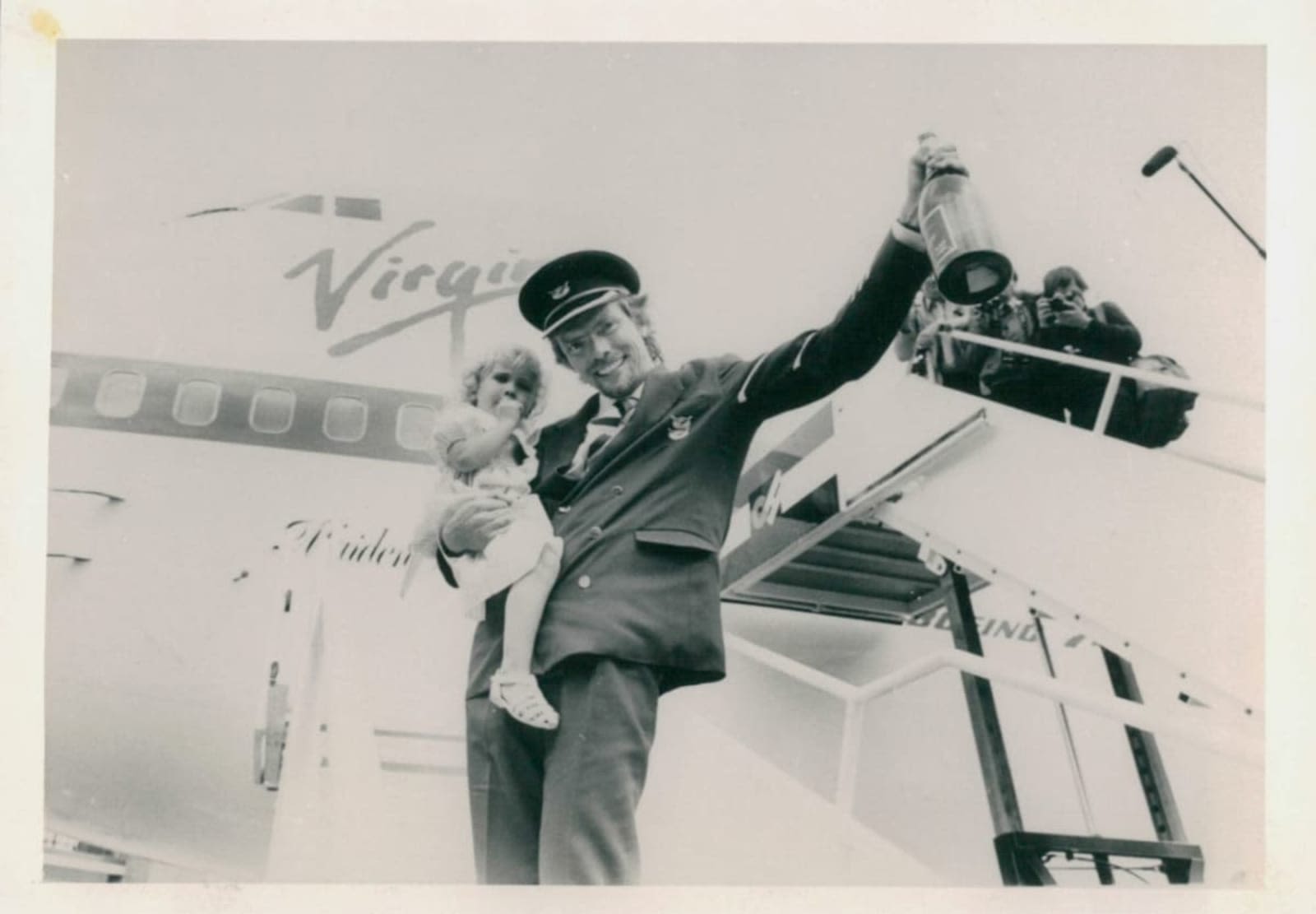 Black and white image of Richard Branson dressed as a pilot, holding Holly and a bottle of champagne, in front of a Virgin aeroplane