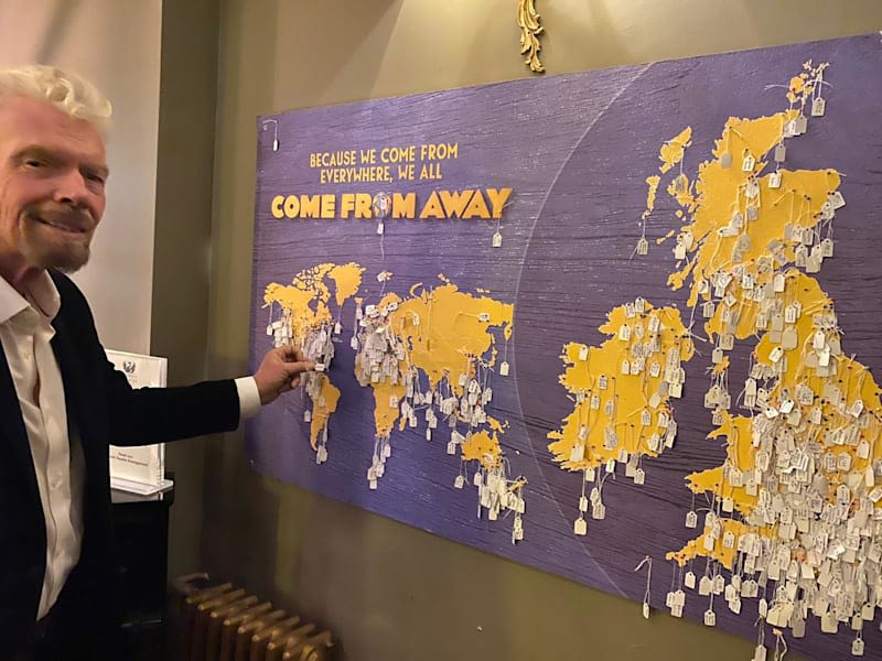 Richard Branson in front of a board that reads 'Because we come from everywhere, we all come from away' with the world map and several pinned pieces of paper 