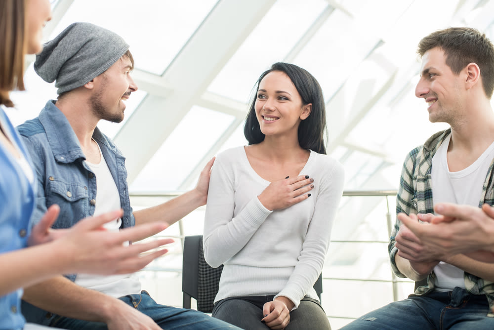 Four people sit talking, one woman has her hand on her chest and a man has his hand on her shoulder