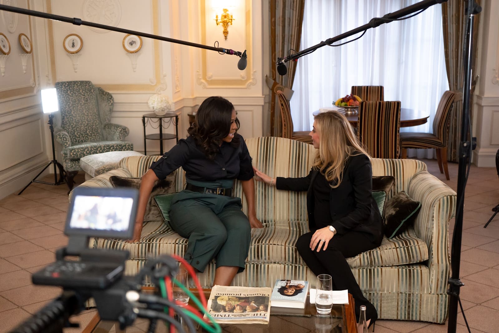 Michelle Obama and Holly Branson sit on a sofa chatting together. Filming equipment is also in shot