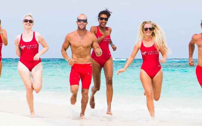 Five people wearing red swimsuits or swim shorts run on a white sandy beach