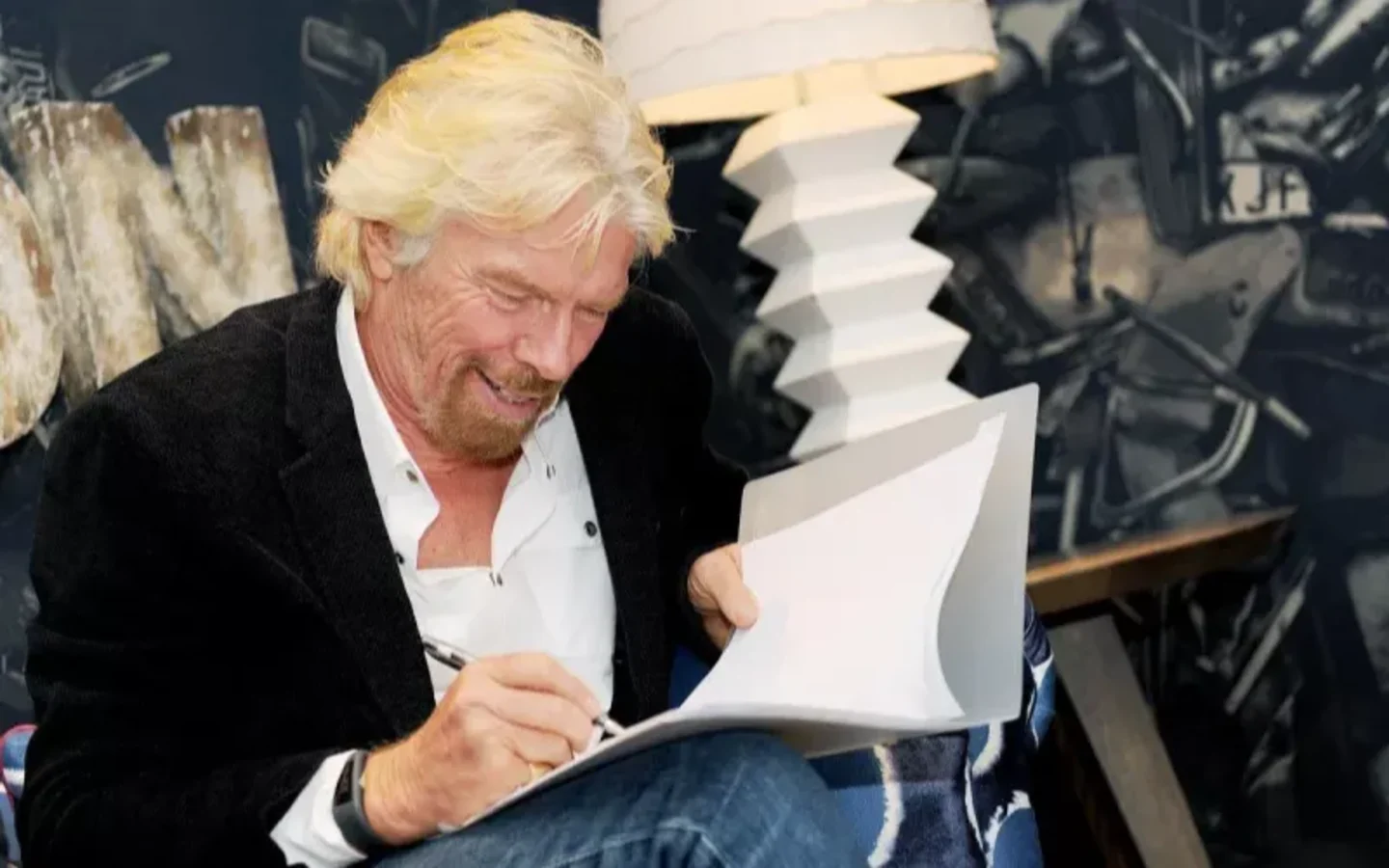 Richard Branson sitting in a chair writing and smiling 