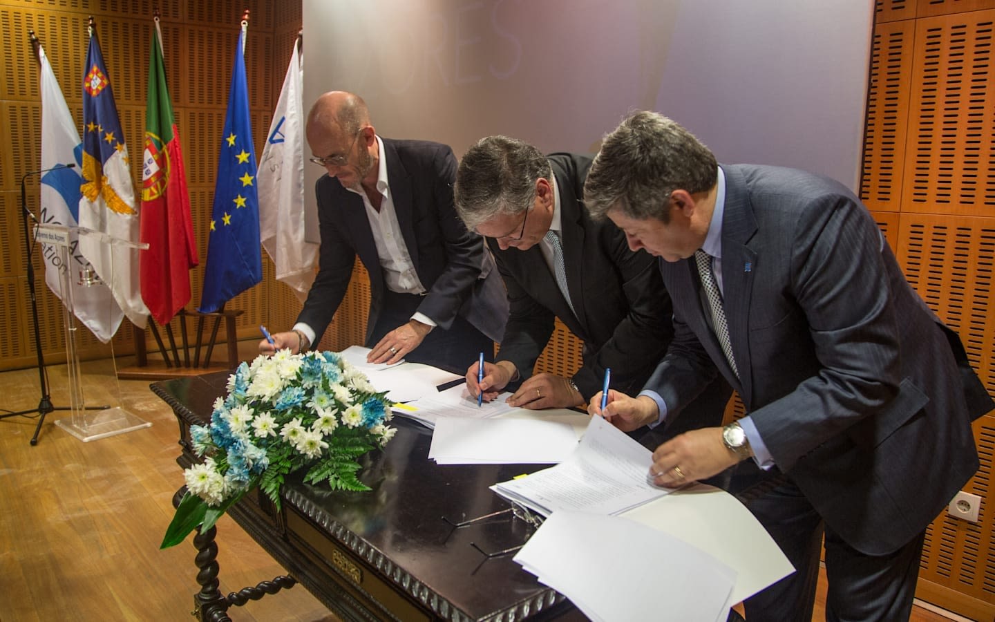 3 politictians stood over a a desk signing papers flags in the back ground and boquet of white and blue flowers on the desk
