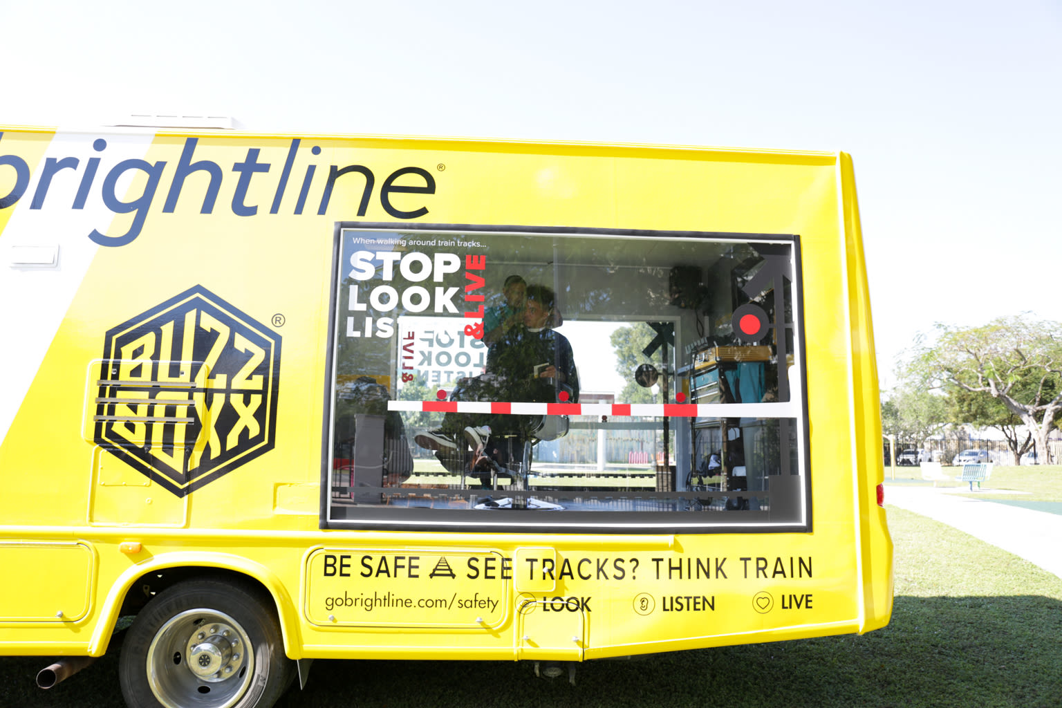 A bright yellow bus with a large window on the side through which you can see two people. Text on the bus says 'be safe. see tracks? Think train.'