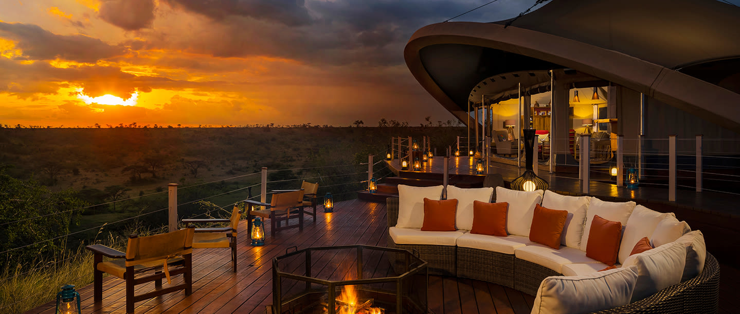 An orange sunset in the distance, in the foreground, outdoor seating surrounds a small firepit