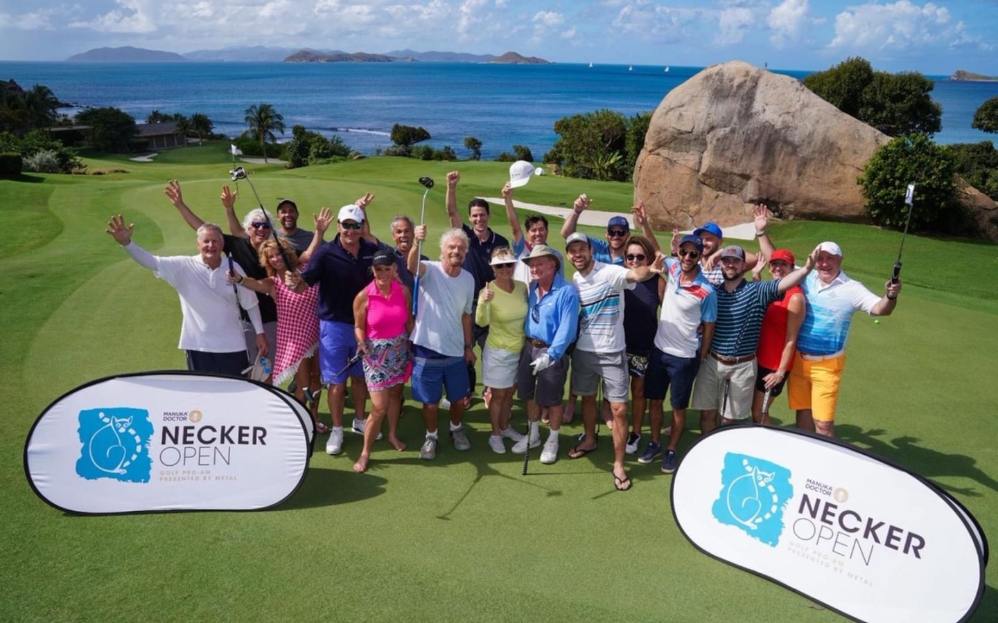 Richard Branson stands with a group of golfers at the Necker Open.  It's a sunny day and the sea is visible in the distance behind them