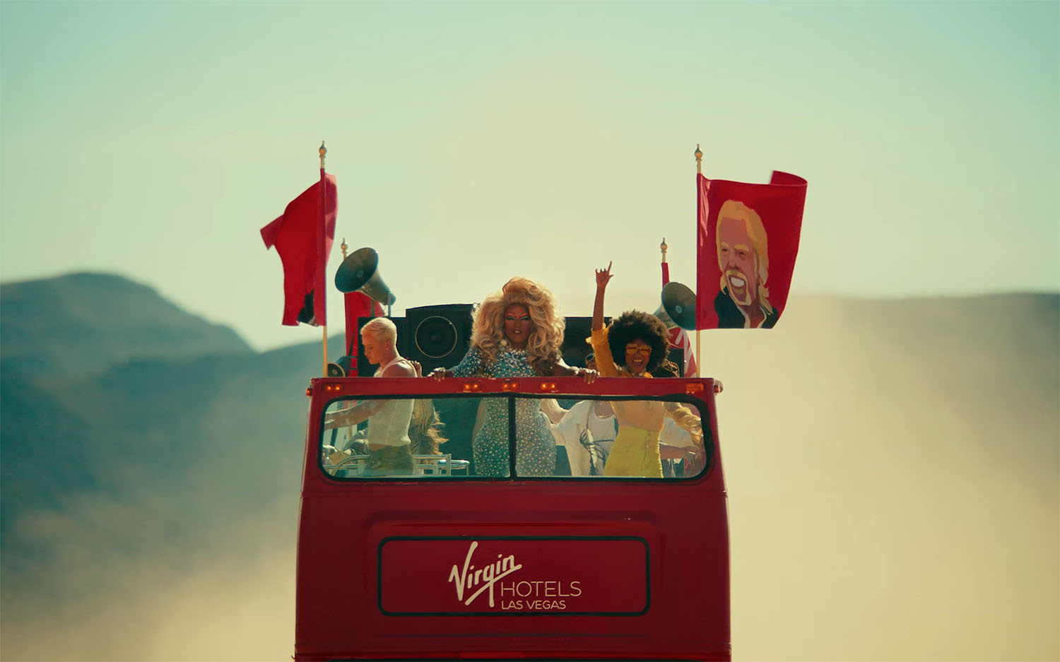 An open top red bus driving through the Las Vegas desert with people partying on the top deck and flags with Richard Branson's face on. The destination on the bus reads: Virgin Hotels Las Vegas