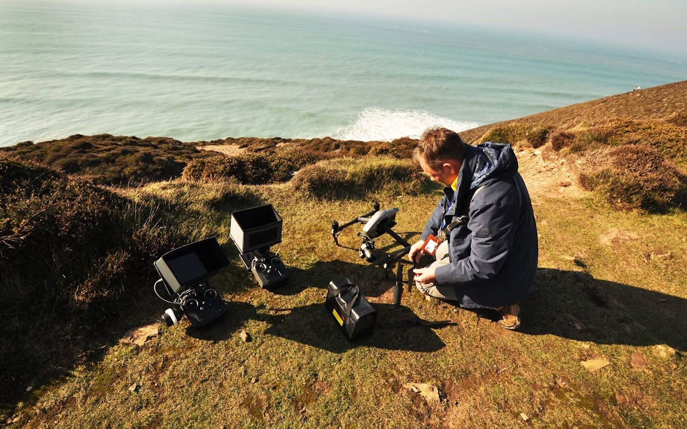 Adrian McDowell crouched next to drones on top of a cliff with sea views