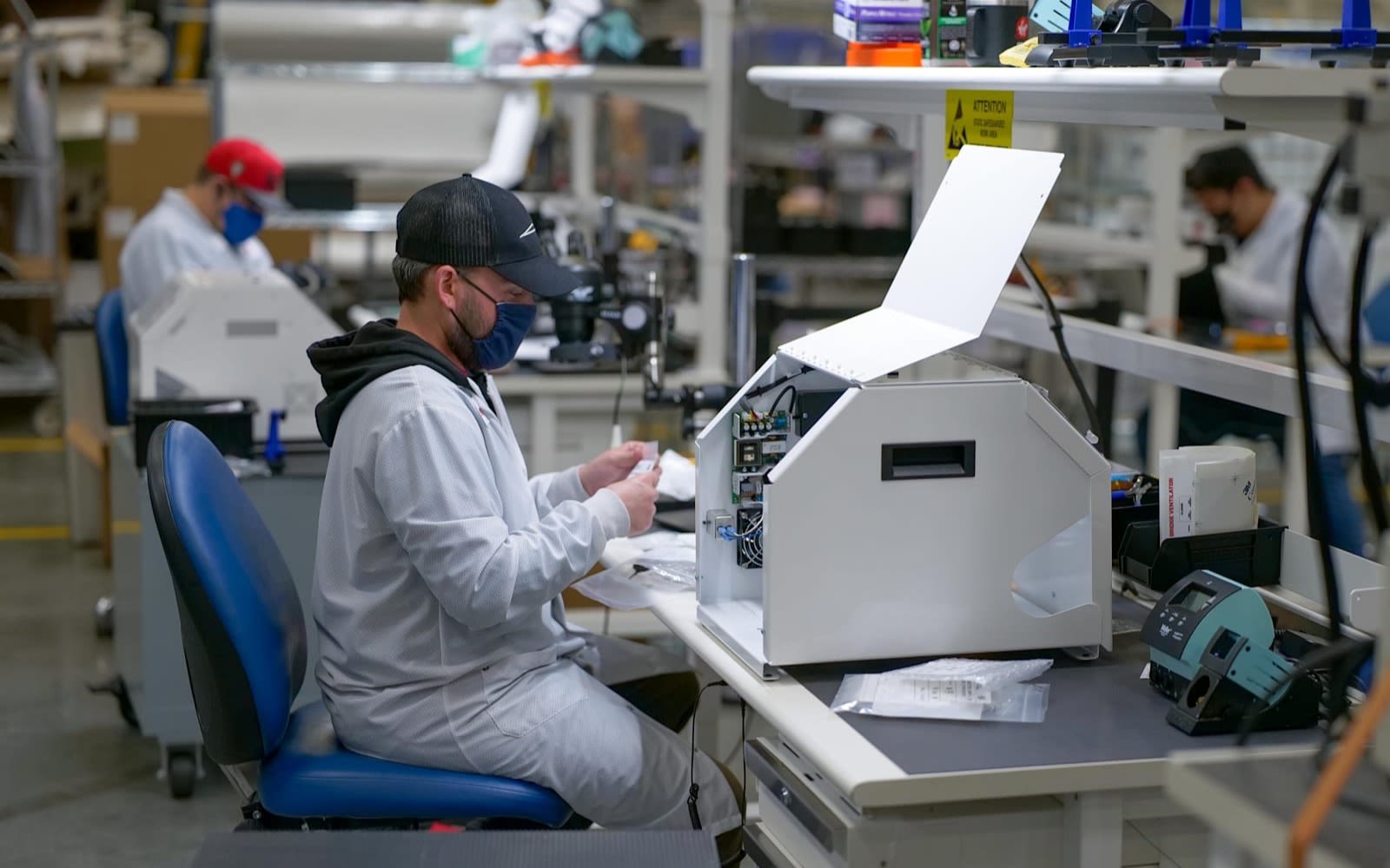 picture showing 3 people at work stations working on medical kit wearing masks