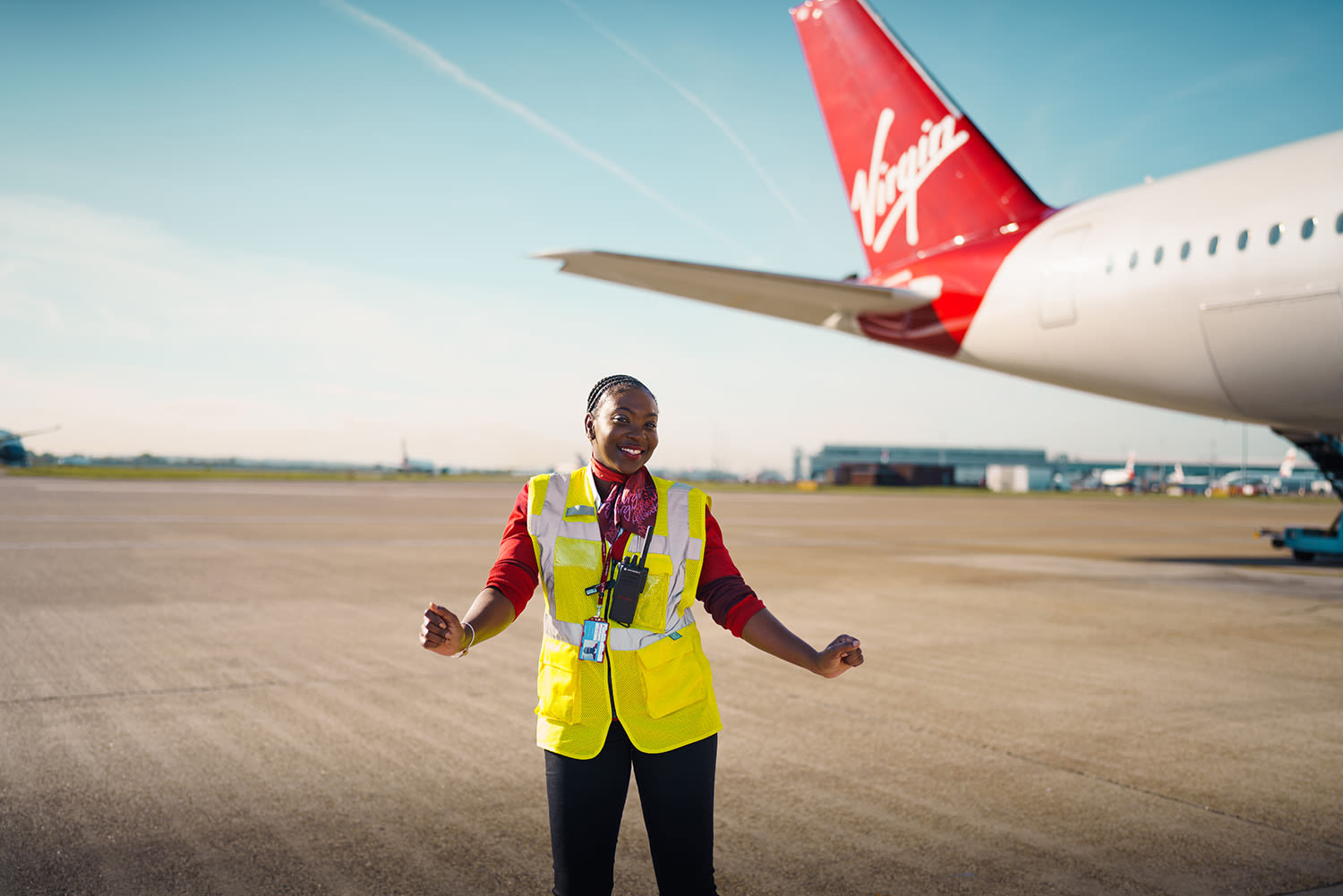 A black woman dancing on a runway wearing a Virgin Atlantic uniform and high-vis vest with the tail of a Virgin Atlantic in the background