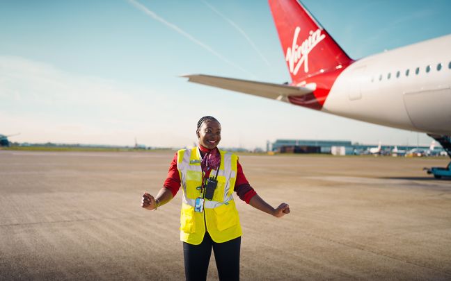 A black woman dancing on a runway wearing a Virgin Atlantic uniform and high-vis vest with the tail of a Virgin Atlantic in the background