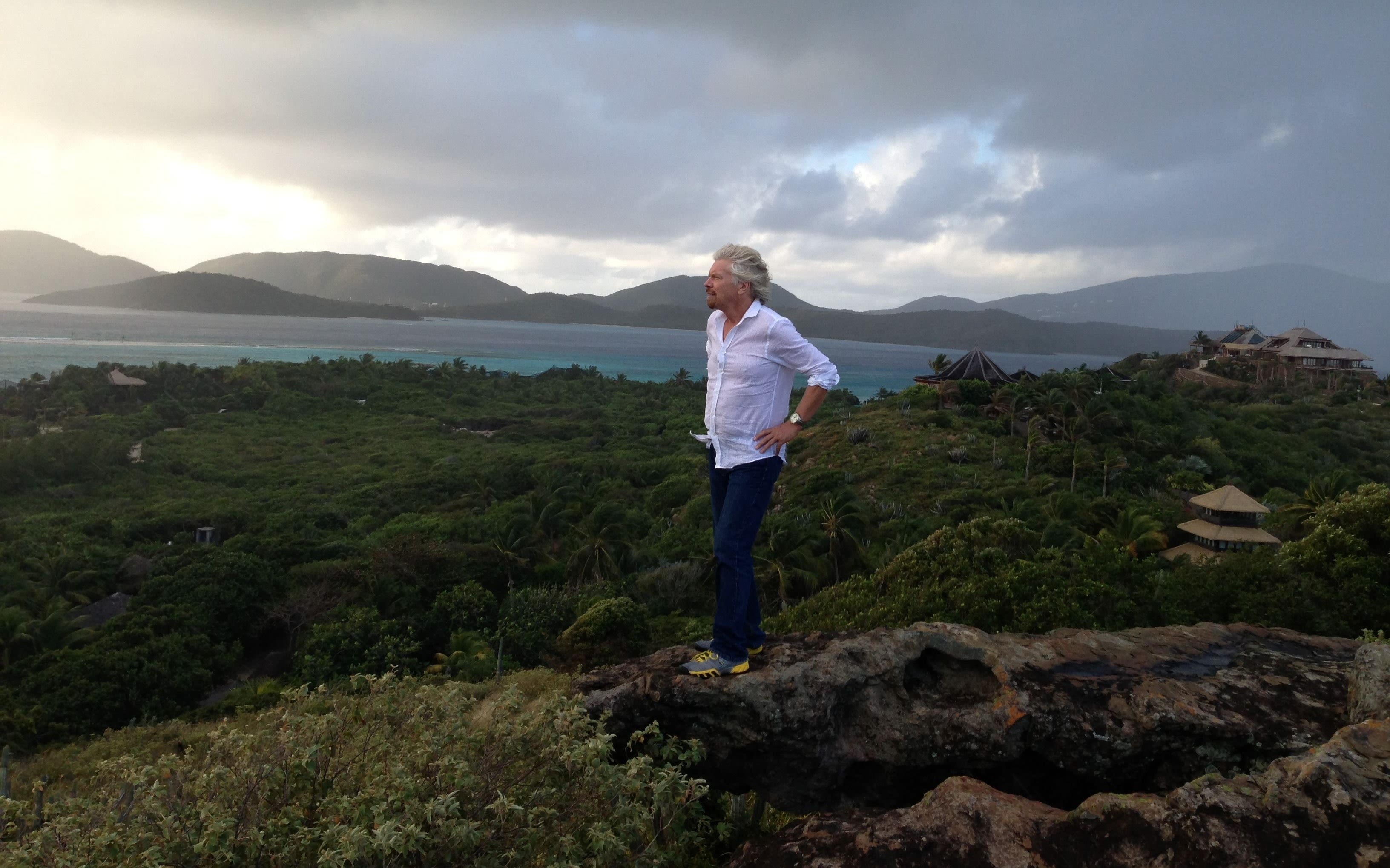 Richard Branson standing on a hill on Necker Island with the house and the ocean in the background