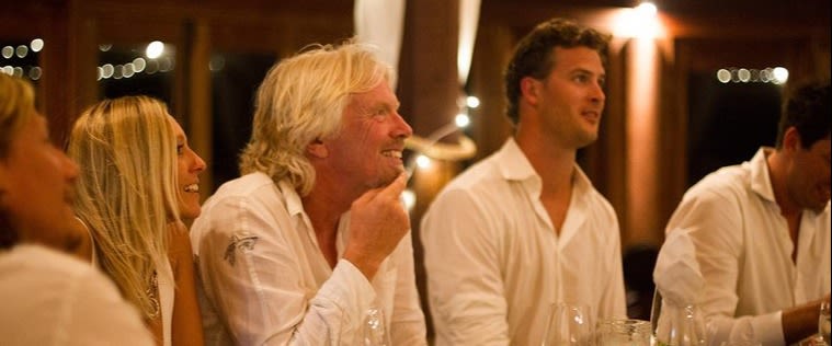 Richard Branson sitting at a table with four others smiling and listening to someone speaking who is out of shot