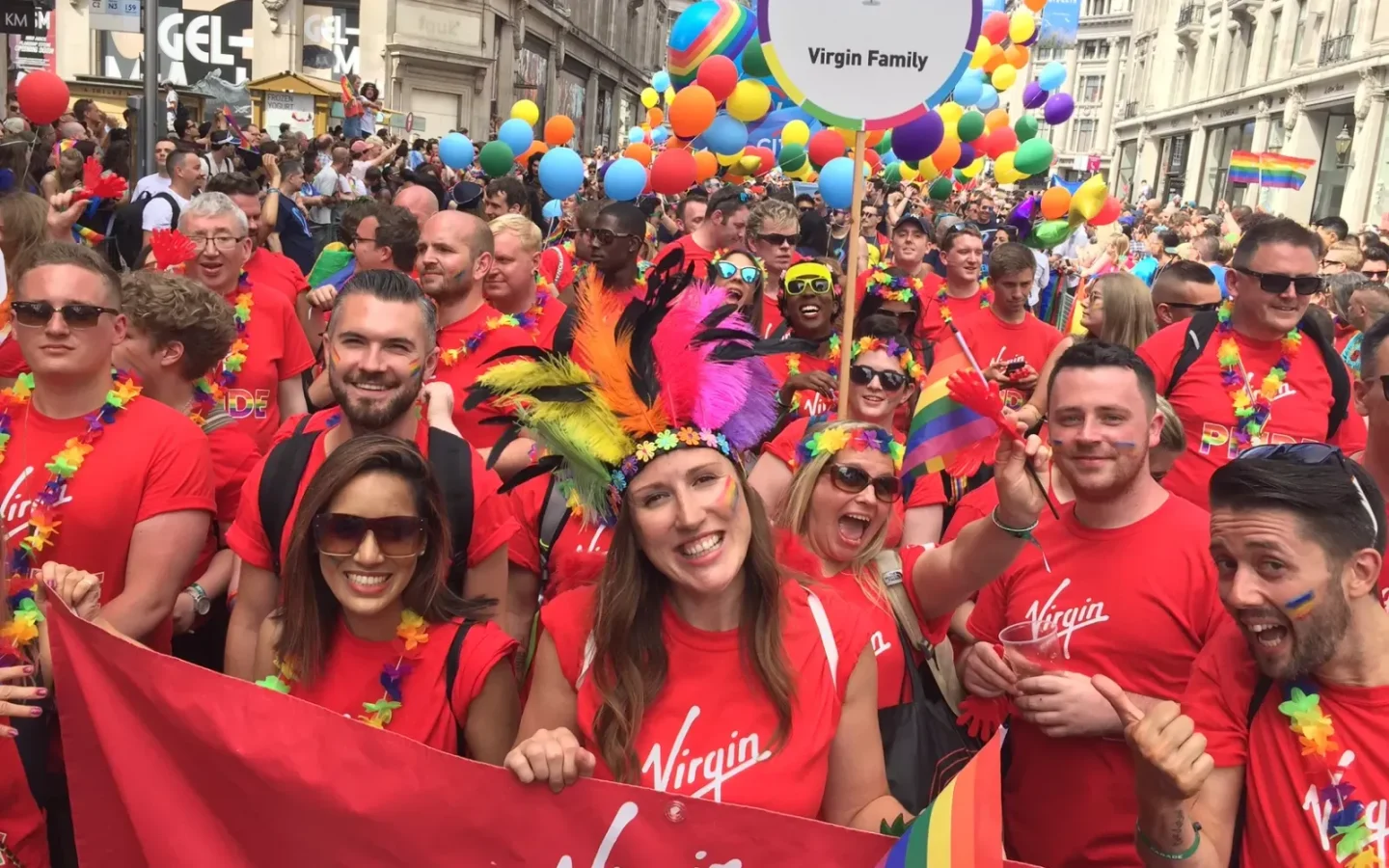 A group of people wearing red tshirts with the Virgin logo on them during the Pride parade. There are lots of rainbow flags and balloons around them