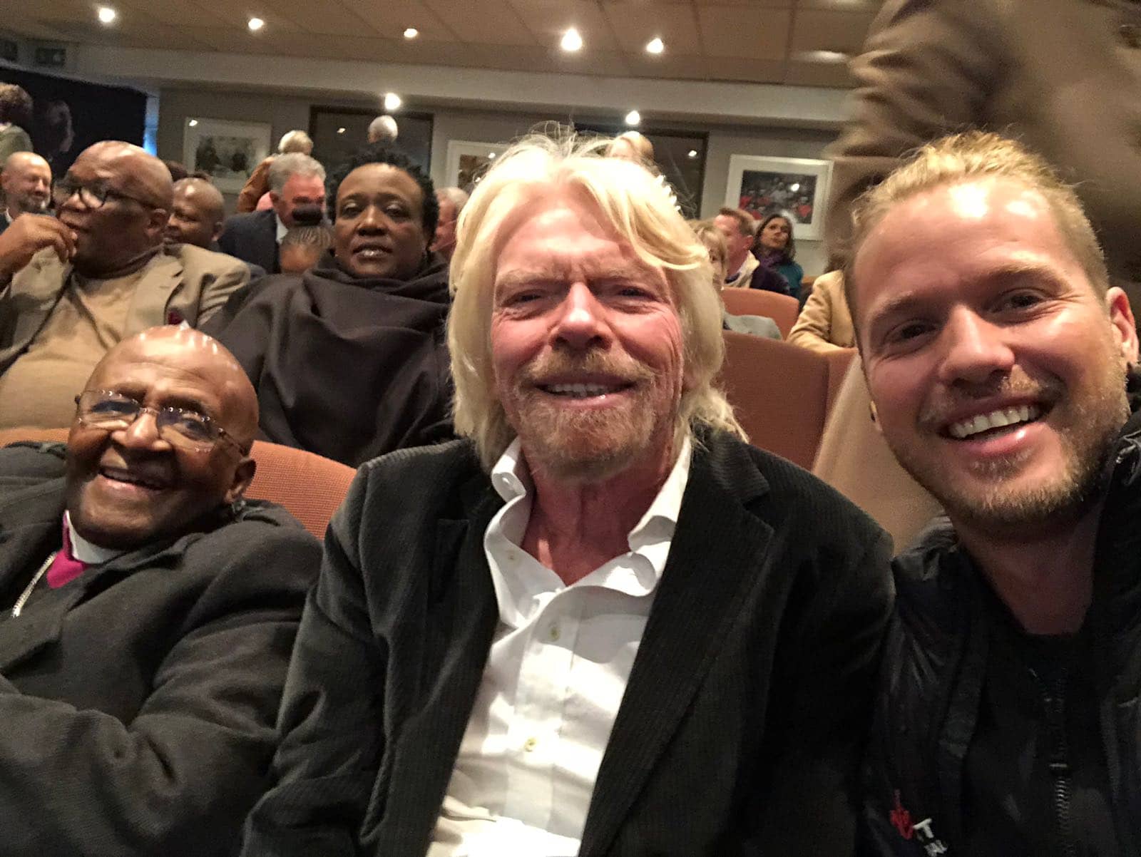 Richard and Sam Branson sitting next to Archbishop Desmond Tutu in a hall with other people in the background 