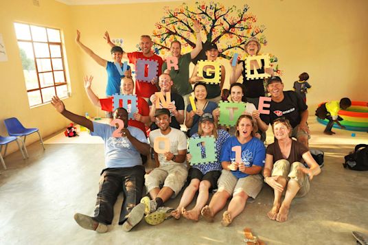 A group of people stand and sit in front of a tree mural on a yellow wall, each holding a letter or number to spell out 'Virgin Unite 2011'.