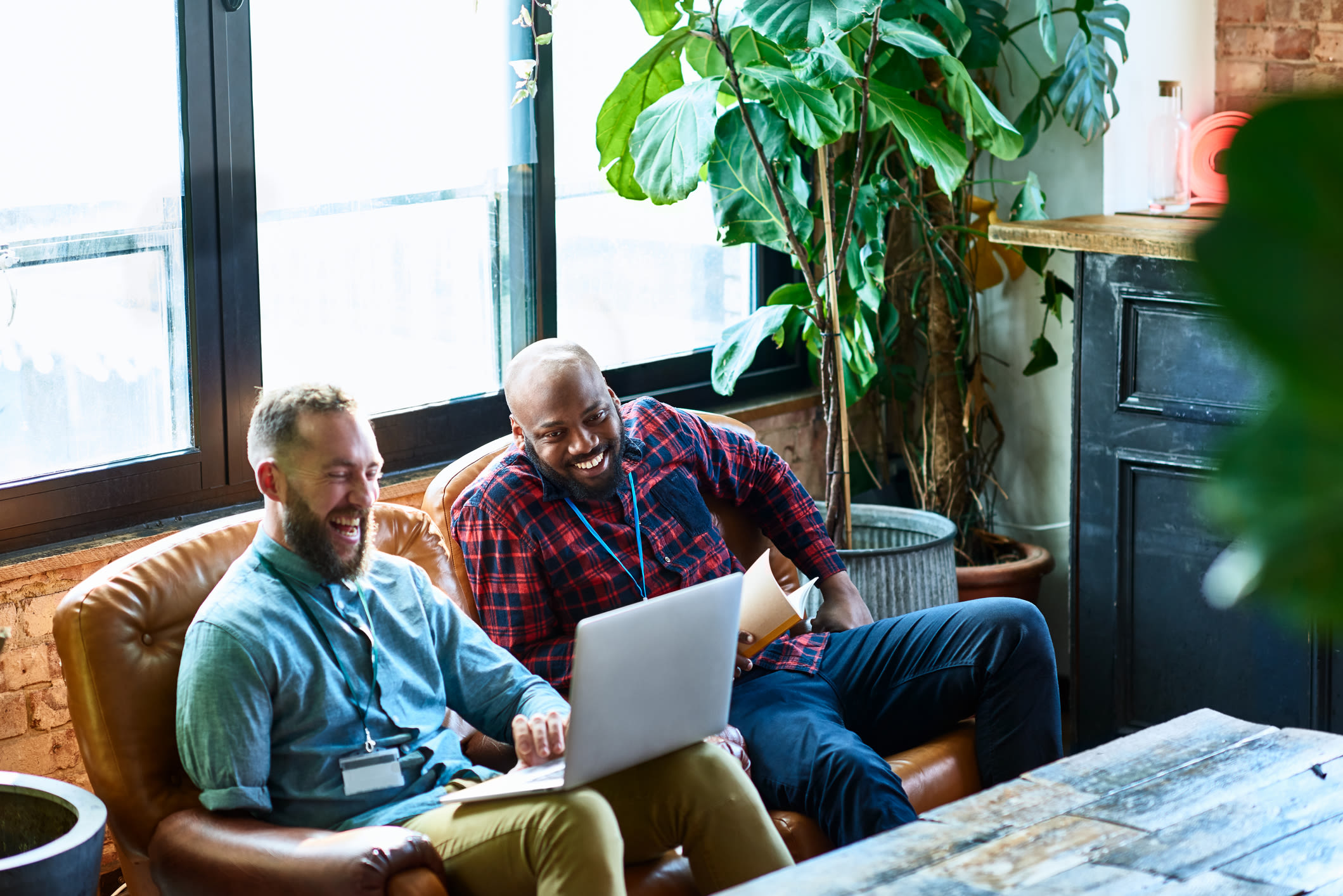 Two men sit on a sofa looking and smiling at a laptop screen