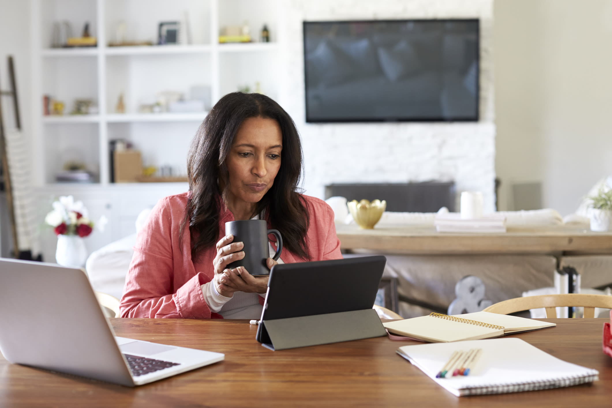 A woman working at home, holding a mug and looking at a tablet