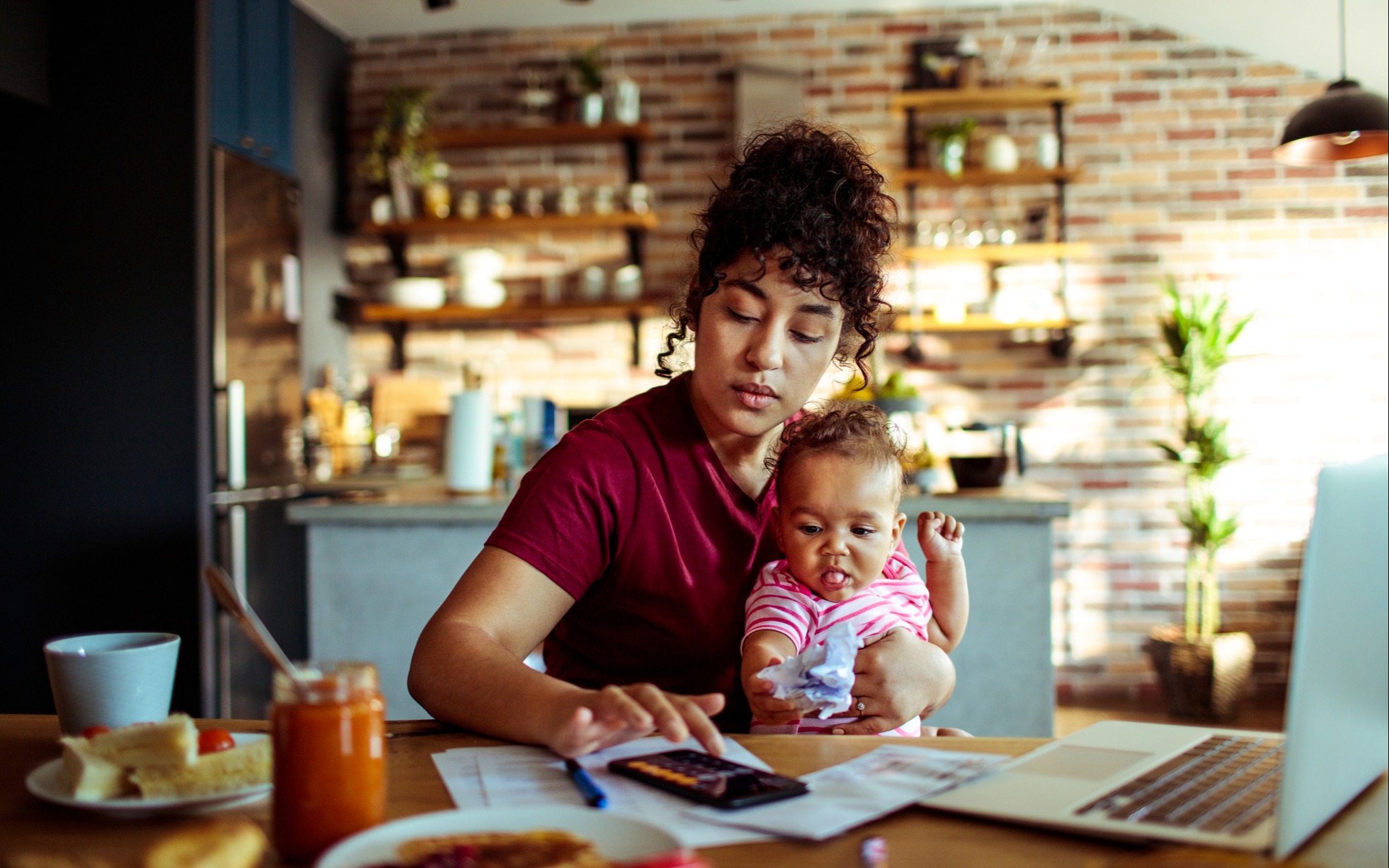 A mum tries to work at a kitchen table with a young child on her lap