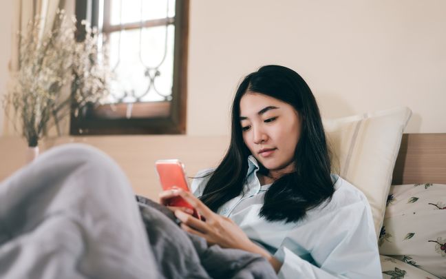 A young woman sitting on her bed looking at her phone